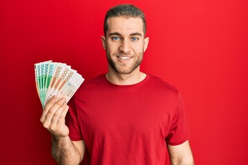 Young caucasian man holding 5000 south korean won banknotes looking positive and happy standing and smiling with a confident smile showing teeth