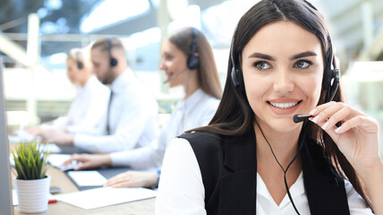 Female customer support operator with headset and smiling, with collegues at background.