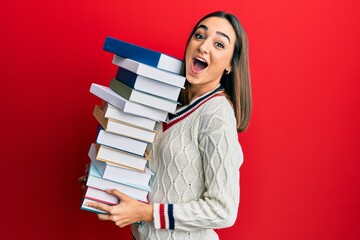 Young brunette student girl holding a pile of books afraid and shocked with surprise and amazed expression, fear and excited face.