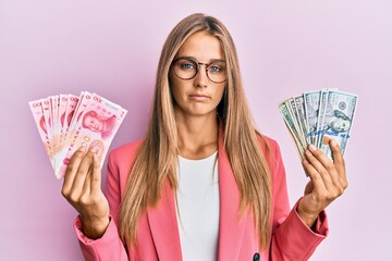Young blonde woman wearing business style holding yuan chinese banknotes and dollars relaxed with serious expression on face. simple and natural looking at the camera.