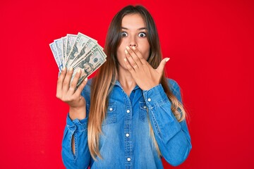Beautiful caucasian woman holding dollars covering mouth with hand, shocked and afraid for mistake....