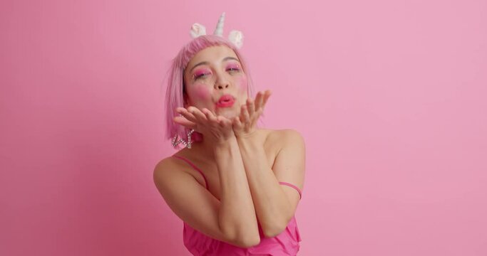 Pretty pink haired Asian girl blows air kiss with affectionate romantic expression makes victory gesture expresses love to you wears dress poses against rosy background. People body language concept.