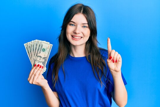 Young beautiful caucasian girl holding 20 dollars banknote smiling with an idea or question pointing finger with happy face, number one