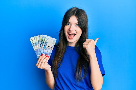 Young beautiful caucasian girl holding south african 100 rand banknotes pointing thumb up to the side smiling happy with open mouth