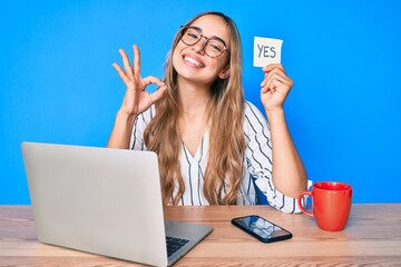 Young beautiful blonde woman holding yes reminder sitting on the table doing ok sign with fingers,...