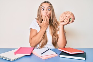 Young beautiful blonde woman holding brain while studying for school covering mouth with hand,...