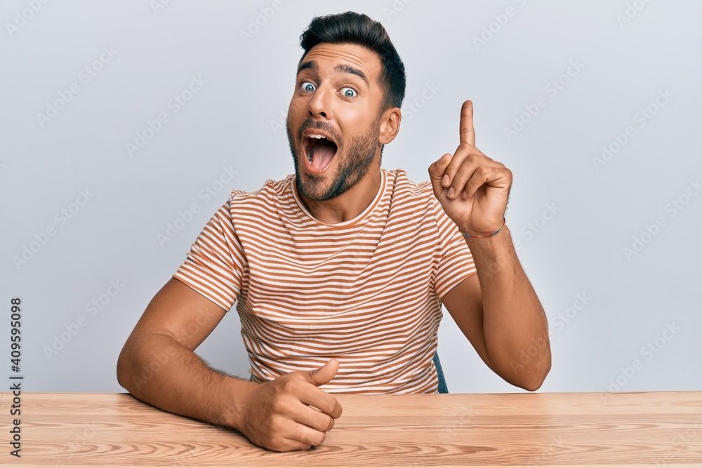 Wall mural handsome hispanic man wearing casual clothes sitting on the table pointing finger up with successful