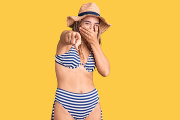 Young beautiful blonde woman wearing bikini and hat laughing at you, pointing finger to the camera with hand over mouth, shame expression