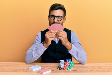 Handsome hispanic croupier man sitting on the table with poker chips and cards winking looking at...