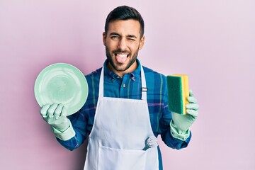 Young hispanic man wearing apron holding scourer washing dishes sticking tongue out happy with...