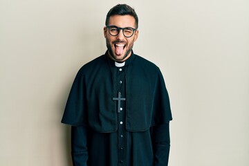 Young hispanic man wearing priest uniform standing over white background sticking tongue out happy with funny expression. emotion concept.