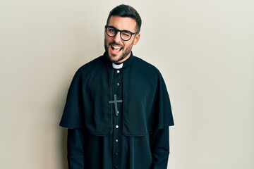 Young hispanic man wearing priest uniform standing over white background winking looking at the...