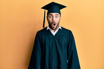 Young hispanic man wearing graduation cap and ceremony robe afraid and shocked with surprise...