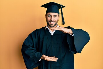 Young hispanic man wearing graduation cap and ceremony robe gesturing with hands showing big and large size sign, measure symbol. smiling looking at the camera. measuring concept.