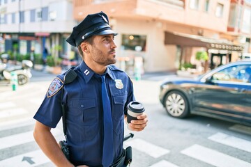 Young hispanic policeman wearing police uniform with serious expression. Drinking cup of take away coffee standing at town street.