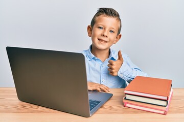 Adorable caucasian kid studying for school exam using laptop smiling happy and positive, thumb up doing excellent and approval sign