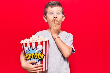 Cute blond kid holding popcorn covering mouth with hand, shocked and afraid for mistake. surprised expression