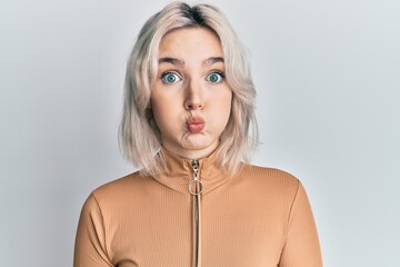 Young blonde girl wearing casual clothes puffing cheeks with funny face. mouth inflated with air,...