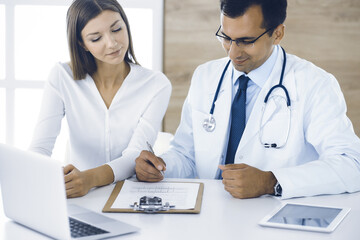 Doctor and patient discussing medical exam results while sitting at the desk in clinic. Male physician using clipboard for filling up medication history record of young woman. Data in medicine