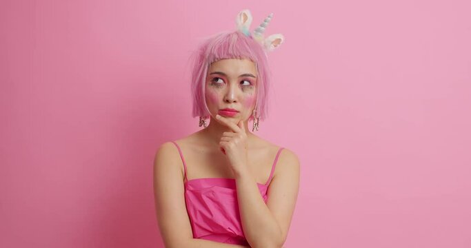 Dissatisfied Asian woman has spoiled makeup after crying looks at different sides thoughtfully tries to decide how to solve problem wears festive dress and unicorn headband isolated on pink background