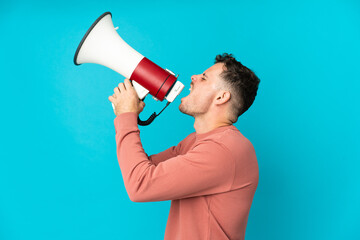Young caucasian handsome man isolated on blue background shouting through a megaphone to announce something in lateral position