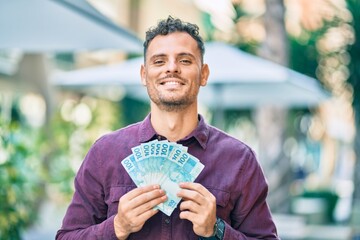Young hispanic man smiling happy holding brazilian real banknotes at the city.