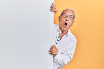Senior caucasian man holding blank empty banner scared and amazed with open mouth for surprise,...