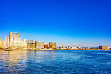 Obraz premium 青空を背景に対岸沿いに建つホテルやビル等の建物見た静かな朝の海の風景