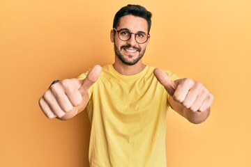 Fototapeta premium Young hispanic man wearing casual clothes and glasses approving doing positive gesture with hand, thumbs up smiling and happy for success. winner gesture.