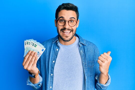 Young hispanic man holding bunch of 20 euro banknotes pointing thumb up to the side smiling happy with open mouth