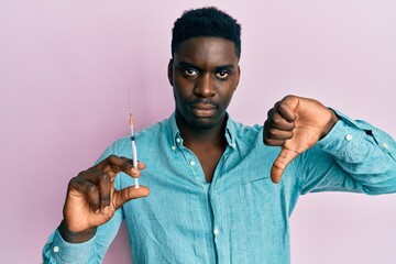 Handsome black man holding syringe with angry face, negative sign showing dislike with thumbs down,...