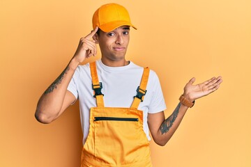 Young handsome african american man wearing handyman uniform over yellow background confused and annoyed with open palm showing copy space and pointing finger to forehead. think about it.