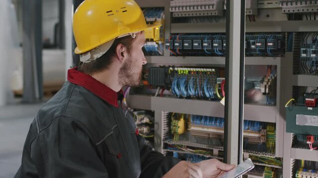 Tilting-up slowmo close-up of electrician in safety helmet installing electric panel at plant with digital tablet