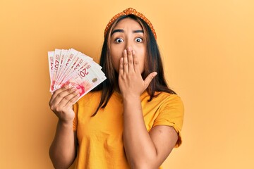 Young latin woman holding israel shekel banknotes covering mouth with hand, shocked and afraid for...