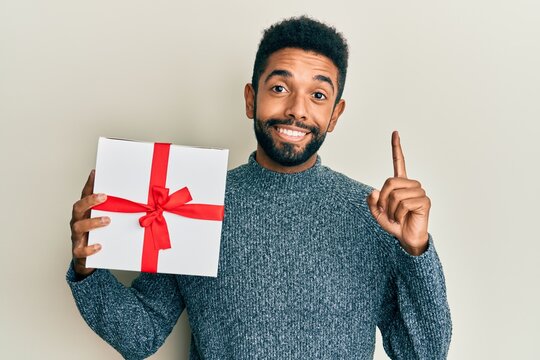 Handsome hispanic man with beard holding gift smiling with an idea or question pointing finger with happy face, number one