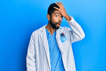 Handsome hispanic man with beard wearing doctor uniform surprised with hand on head for mistake,...