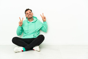 Young handsome caucasian man sitting on the floor showing victory sign with both hands