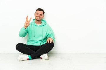 Young handsome caucasian man sitting on the floor happy and counting three with fingers