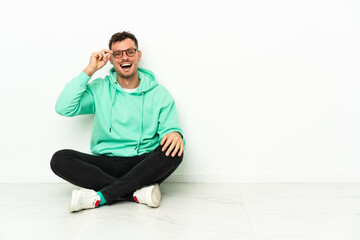 Young handsome caucasian man sitting on the floor with glasses and surprised