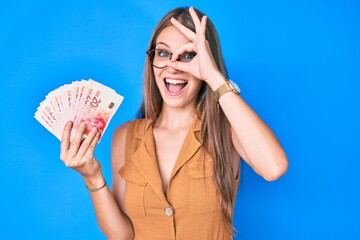 Young blonde girl holding israeli shekels smiling happy doing ok sign with hand on eye looking...