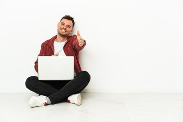 Young handsome caucasian man sit-in on the floor with laptop shaking hands for closing a good deal