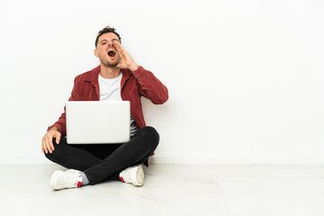 Young handsome caucasian man sit-in on the floor with laptop shouting with mouth wide open