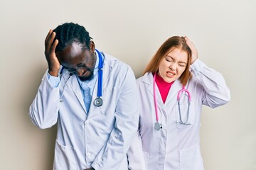 Young interracial couple wearing doctor uniform and stethoscope suffering from headache desperate and stressed because pain and migraine. hands on head.