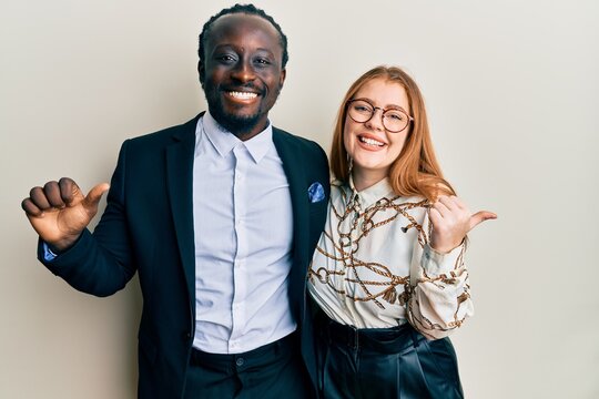 Young interracial couple wearing business and elegant clothes pointing to the back behind with hand and thumbs up, smiling confident