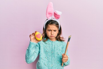 Little beautiful girl wearing cute easter bunny ears holding colored egg skeptic and nervous, frowning upset because of problem. negative person.