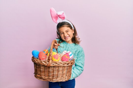 Little beautiful girl wearing cute easter bunny ears holding wicker basket with colored eggs winking looking at the camera with sexy expression, cheerful and happy face.