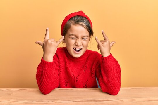 Little beautiful girl wearing casual clothes sitting on the table shouting with crazy expression doing rock symbol with hands up. music star. heavy concept.