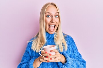 Young blonde girl wearing winter sweater and drinking a cup of hot coffee celebrating crazy and...