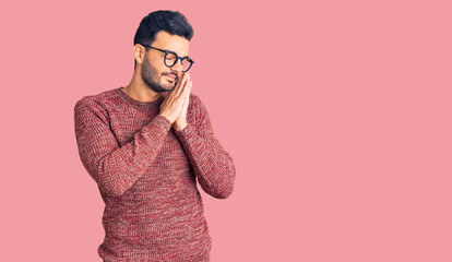Young handsome hispanic man wearing winter sweater and glasses sleeping tired dreaming and posing with hands together while smiling with closed eyes.