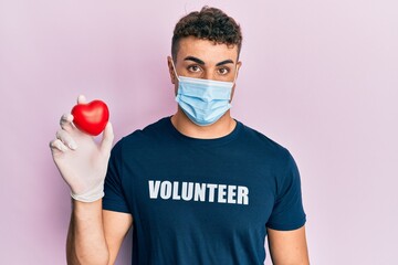 Hispanic young man wearing medical mask wearing volunteer t shirt holding heart skeptic and...